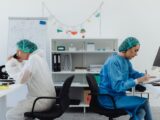 Two scientists in a lab, one examining a microscope, the other taking notes.