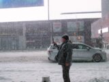 Man stands on snowy street with cars and buildings.