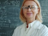 Woman in lab coat and glasses in front of blackboard.