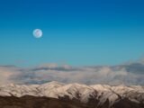 Capture of snow-covered mountains under a blue sky with the moon in view, showcasing nature's serene beauty.