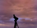 Silhouette of a telescope on a tripod set against a vibrant sunset sky with clouds.