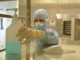 A scientist in protective gear reaching for a sanitizer bottle in a laboratory setting.